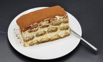 A piece of tiramisu on a white plate. The dessert is dusted with cocoa and has a layered structure.