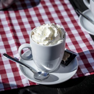 A cup with cream on a white saucer. The background is a red and white checked table runner. | © Kleinwalsertal Tourismus | Dominik Berchtold