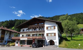 A charming building in alpine style with various balconies. In the background, green hills and a blue sky can be seen. | © Kleinwalsertal Tourismus | Nicole Lughammer