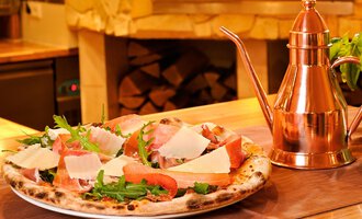 A freshly baked pizza with ham, arugula, and Parmesan. Next to the pizza is an elegant, copper oil container. | © Ristorante Trentino | Michael Monschau