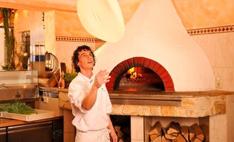 A pizza maker tosses a pizza dough into the air. In the background, a traditional wood-fired oven can be seen. | © Ristorante Trentino | Michael Monschau