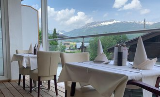 An elegant restaurant area with white tables and chairs. In the background, green mountains and a blue sky can be seen. | © Ristorante Trentino | Dietmar Jochum