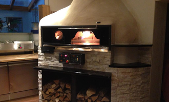 A modern wood stove in a kitchen with a rustic stone oven cart. The stove has a large opening and wood stored underneath. | © Ristorante Trentino | Dietmar Jochum