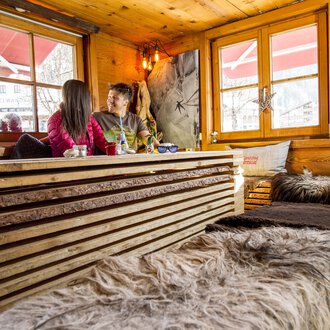 A cozy café with wooden furniture and soft blankets. Two people are sitting by the window and enjoying their time together. | © Kleinwalsertal Tourismus | Dominik Berchtold