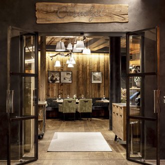 An inviting entrance to a restaurant with wooden details and warm lighting. The atmosphere is cozy and welcoming. | © Travel Charme Ifen Hotel