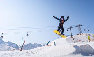 A snowboarder jumps over a snow hill in front of a clear mountain landscape. In the background, ski lifts and other skiers can be seen. | © OBERSTDORF · KLEINWALSERTAL BERGBAHNEN