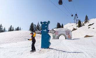 A skier stands on a snow-white slope next to a blue bear and an architectural element. In the background, lifts and snow-covered trees can be seen. | © OBERSTDORF · KLEINWALSERTAL BERGBAHNEN