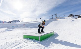 A snowboarder jumps over a green box in the snow. In the background, mountains and ski lifts are visible. | © OBERSTDORF · KLEINWALSERTAL BERGBAHNEN