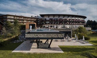 Ein modernes Hotel mit einem großen, gläsernen Pool steht im Vordergrund. Die umliegende Landschaft ist grün und einladend. | © Travel Charme Ifen Hotel