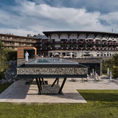 Ein modernes Hotel mit einem großen, gläsernen Pool steht im Vordergrund. Die umliegende Landschaft ist grün und einladend. | © Travel Charme Ifen Hotel
