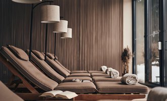Ein entspannender Wellnessbereich mit bequemen Liegen und sanfter Beleuchtung. Große Fenster bieten einen Blick auf die Natur. | © Travel Charme Ifen Hotel