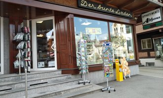 A small shop named "The Colorful Shop" with an inviting display window. In front of the store are sales stands with various items. | © Der Bunte Laden | Romana Volderauer