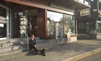 A woman is sitting in front of a store on the street with her dog. The store has large windows and an inviting atmosphere. | © Der Bunte Laden | Romana Volderauer