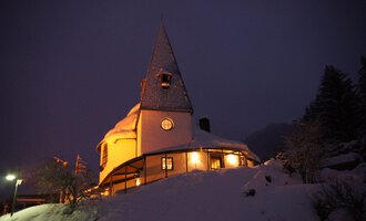 Ein charmantes, schneebedecktes Haus mit einem spitzen Dach in einer winterlichen Landschaft. Die warmen Lichter im Gebäude schaffen eine gemütliche Atmosphäre in der Dunkelheit. | © Ev. Kirche Kleinwalsertal