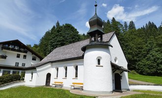 A small, white chapel with a black roof amidst greenery. In the background, trees and a holiday accommodation can be seen. | © Kleinwalsertal Tourismus | N. Lughammer