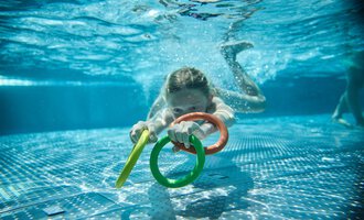 A child swims underwater, holding colorful rings in their hands. The water's surface reflects the light, creating a vibrant atmosphere. | © Kleinwalsertal Tourismus | Oliver Farys