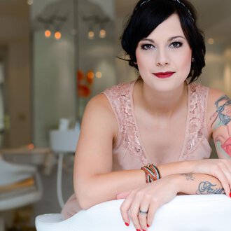 Eine Frau mit Tattoos sitzt lächelnd in einem modernen Raum. Sie trägt ein elegantes, rosafarbenes Kleid und hat einen schicken Look. | © Fräulein Sugar Coiffeur | Sandra Bromm