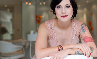 Eine Frau mit Tattoos sitzt lächelnd in einem modernen Raum. Sie trägt ein elegantes, rosafarbenes Kleid und hat einen schicken Look. | © Fräulein Sugar Coiffeur | Sandra Bromm