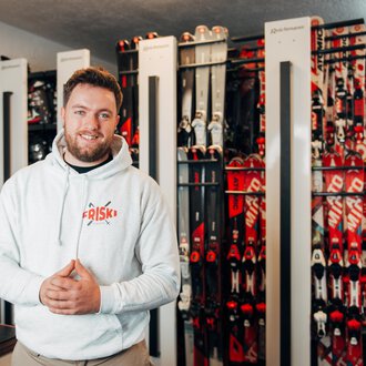 A smiling man stands in a sporting goods store, surrounded by ski equipment. In the background, numerous skis in various colors are lined up. | © Friski Marcellus Fritz | Stefan Klauser