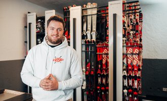 A smiling man stands in a sporting goods store, surrounded by ski equipment. In the background, numerous skis in various colors are lined up. | © Friski Marcellus Fritz | Stefan Klauser