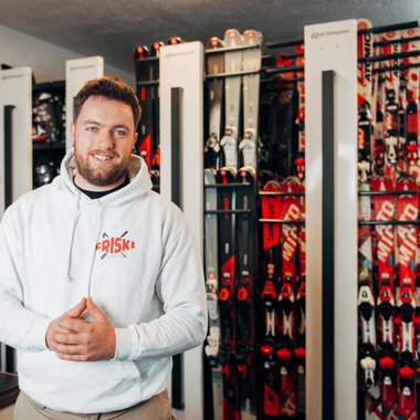 A smiling man stands in a sporting goods store, surrounded by ski equipment. In the background, numerous skis in various colors are lined up. | © Friski Marcellus Fritz | Stefan Klauser