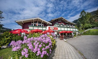 A cozy restaurant with a red facade and blooming flowers in the foreground. Red umbrellas provide shade for the guests. | © Gasthof Alpenblick | Theodor Pinn