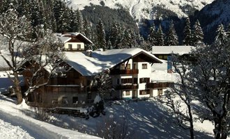 A picturesque winter landscape with snow-covered mountains in the background. In the foreground stands a charming house, surrounded by snow-covered trees. | © Gasthof Café Alpenwald | Thorsten Alpenwald