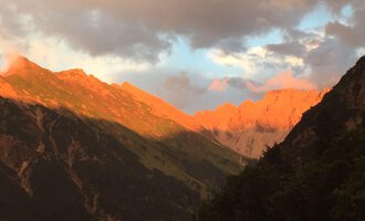 An impressive mountain landscape with a sunset that illuminates the rocks in warm orange tones. The sky is cloudy, creating a dramatic atmosphere. | © Gasthof Café Alpenwald | Thorsten Alpenwald