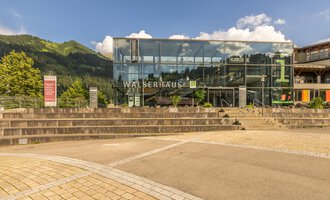 Ein modernes Gebäude mit großer Glasfassade, das Walserhaus heißt. Es liegt in einer malerischen Berglandschaft. | © Kleinwalsertal Tourismus | Steffen Berschin
