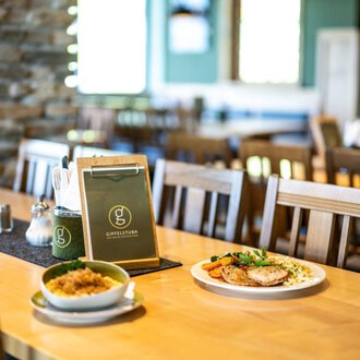 Ein gemütliches Restaurant mit Holzstühlen und einem sauberen Tisch. Auf dem Tisch liegt eine Speisekarte, daneben befinden sich ein Teller mit Fleisch und Gemüse sowie eine Schüssel mit Reis. | © OBERSTDORF·KLEINWALSERTAL BERGBAHNEN