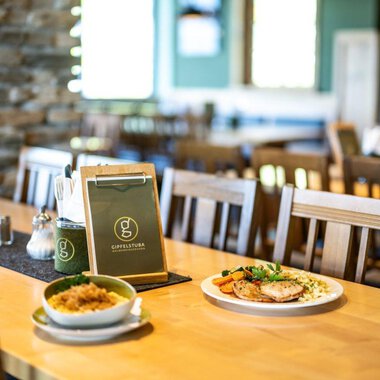 A cozy restaurant with wooden chairs and a clean table. On the table lies a menu, next to it is a plate with meat and vegetables as well as a bowl of rice. | © OBERSTDORF·KLEINWALSERTAL BERGBAHNEN