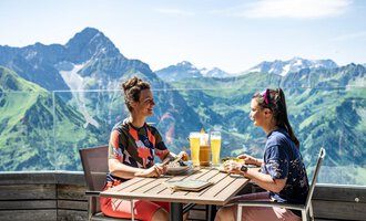 Zwei Frauen sitzen an einem Tisch und genießen Getränke und Essen. Im Hintergrund sind grüne Berge und ein blauer Himmel sichtbar. | © Oberstdorf · KLEINWALSERTAL BERGBAHNEN