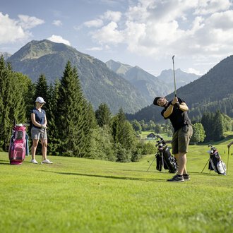 A group of golfers is practicing on a picturesque golf course with mountains in the background. The sun is shining, and the landscape is green and inviting. | © Golfclub Oberstdorf | ©kunst.joachimweiler.de