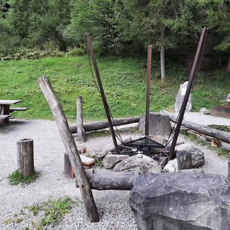Ein gemütlicher Grillplatz im Freien mit einem großen Feuerkreis aus Steinen und Holz. Im Hintergrund steht eine Picknickbank auf einer grünen Wiese. | © Kleinwalsertal Tourismus | Veronika Senn