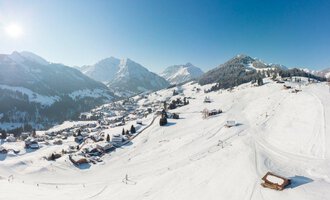 A picturesque winter landscape with snow-covered mountains and a clear blue sky. In the foreground, ski slopes and a small village in the distance can be seen. | © OBERSTDORF · KLEINWALSERTAL BERGBAHNEN