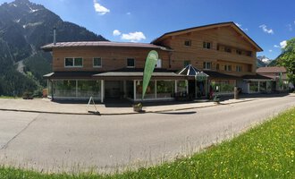 A modern wooden building in the mountains with a clear blue sky. In front of the building, a green meadow stretches out. | © Sport Hilbrand OG