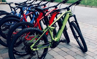 A row of colorful mountain bikes stands on a paved path. In the background, a wooden house and a green meadow can be seen. | © Sport Hilbrand OG