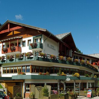 A charming hotel in alpine style with a wavy roof and many balconies. Surrounded by green hills and blue sky. | © IFA Hotel Alpenrose