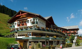 A charming hotel in alpine style with a wavy roof and many balconies. Surrounded by green hills and blue sky. | © IFA Hotel Alpenrose