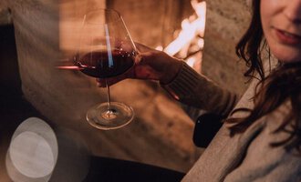 A person is holding a wine glass in their hand. In the background, a fireplace is burning, creating a cozy atmosphere. | © Naturhotel Chesa Valisa | Heldentheater