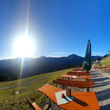 A beautiful mountain landscape with the sun shining over the peaks. In the foreground, there are wooden benches and tables with green sunshades. | © Ifenhütte | Petra Diener