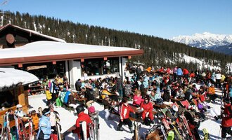 A bustling ski lodge in the mountains, surrounded by snow and trees. Many people are enjoying the sunshine and the wintry atmosphere. | © Ifenhütte | Petra Diener