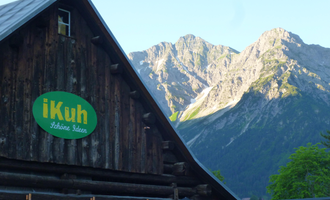 A traditional wooden house with a green sign that says "iKuh." In the background, majestic mountains and a clear sky can be seen. | © iKuh Schöne Ideen | Daniela Neumann