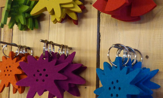 Colorful keychains in the shape of stars hang on a wooden wall. The colors are red, blue, green, yellow, orange, and purple. | © iKuh Schöne Ideen | Daniela Neumann