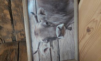 An image of a house with the head of a cow standing in front of a wooden wall. The frame is made of wood and fits the rural surroundings.