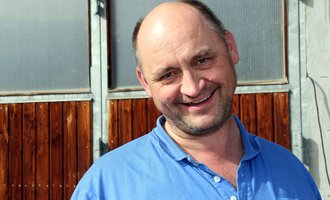 A cheerful man smiles at the camera. Wooden doors and windows can be seen in the background. | © Innere Wiesalpe | Andrea Kainz zuckerschnecke.at
