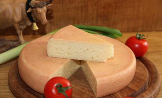 A cheese wheel with a cut slice lies on a wooden board. Around it, fresh tomatoes and a cow figurine can be seen. | © Innere Wiesalpe | Andrea Kainz zuckerschnecke.at