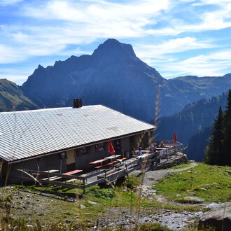 An alpine cabin surrounded by mountains and forests. The sky is clear with a few clouds. | © Kleinwalsertal Tourismus
