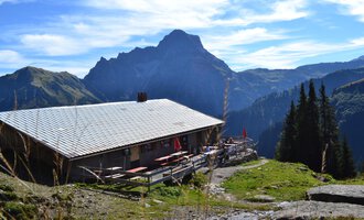 An alpine cabin surrounded by mountains and forests. The sky is clear with a few clouds. | © Kleinwalsertal Tourismus