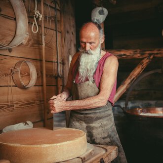 An older man with long white hair and a beard works in a woodworking shop. In the background, a large kettle can be seen. | © Käsestadl Kleinwalsertal | Nadine Paul